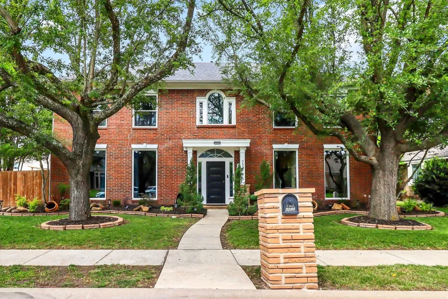 Traditional brick exterior featuring a prominent arched entry, a concrete walkway, mature trees, and a stone-clad mailbox