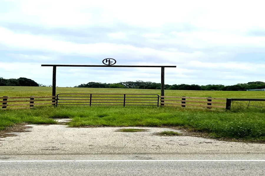Property entrance featuring a metal gate and wooden fence, set against a backdrop of expansive green fields and mature trees
