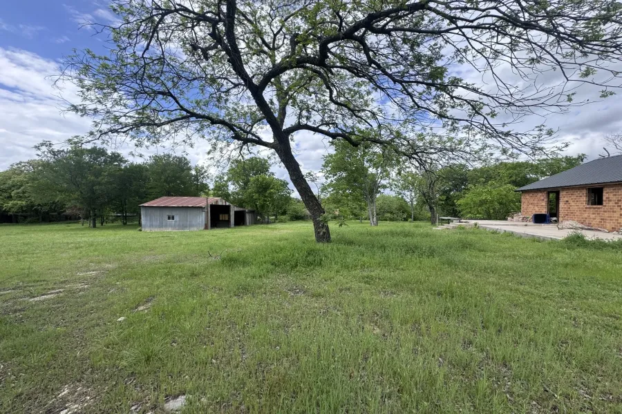 Expansive grassy area featuring a mature tree, an outbuilding with a galvanized metal roof, and a brick structure with a dark shingle roof