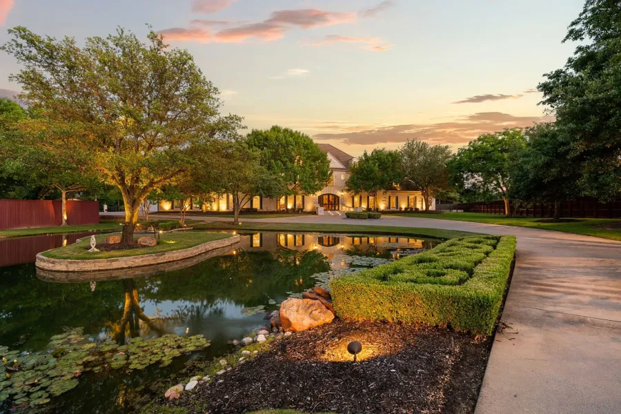 Grand estate facade with illuminated architectural details, circular drive, mature trees, and a private pond featuring a central island